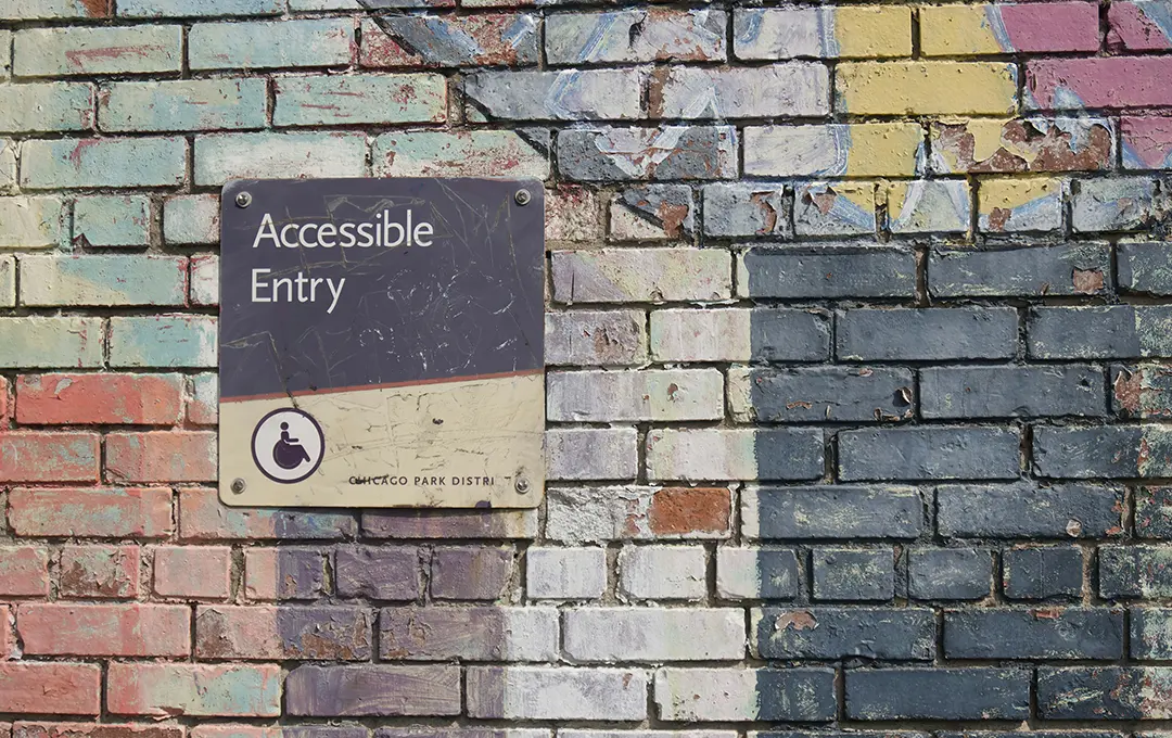 Eine farbenfrohe Backsteinmauer auf der ein Schild mit der Aufschrift „Barrierefreier Eingang“ und ein Rollstuhlsymbol zu sehen sind.